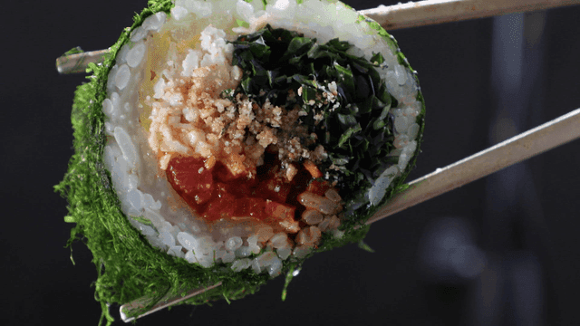 Chopsticks picking up piece of aged kimchi and seaweed roll containing fresh ingredients