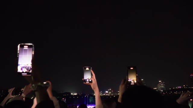 밤에 강변에서 드론 라이트 쇼를 촬영하는 사람들