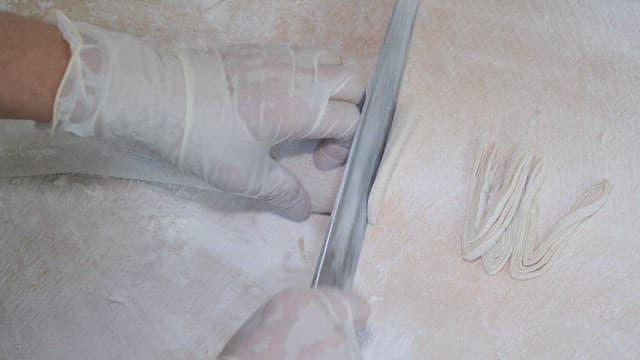 장갑을 낀 사람이 밀가루를 뿌린 표면에서 수제 국수를 자르고 준비하는 모습