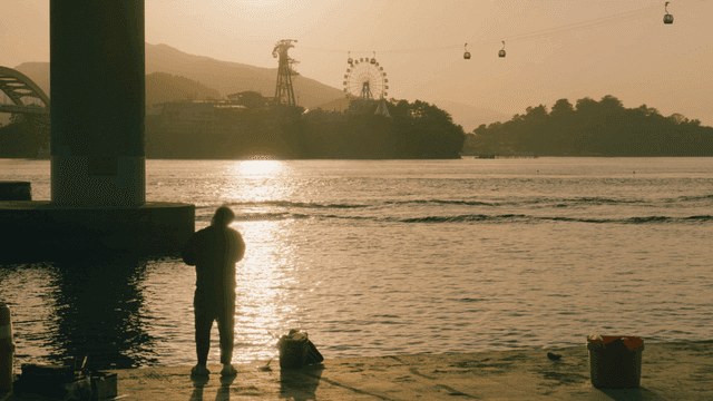 Back of man fishing on riverbank at sunset and cable car
