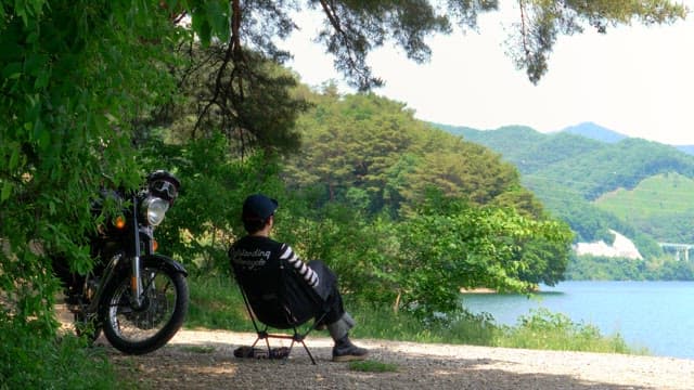 Man relaxing by a lake, sitting on a portable chair under a tree, with a motorcycle parked nearby