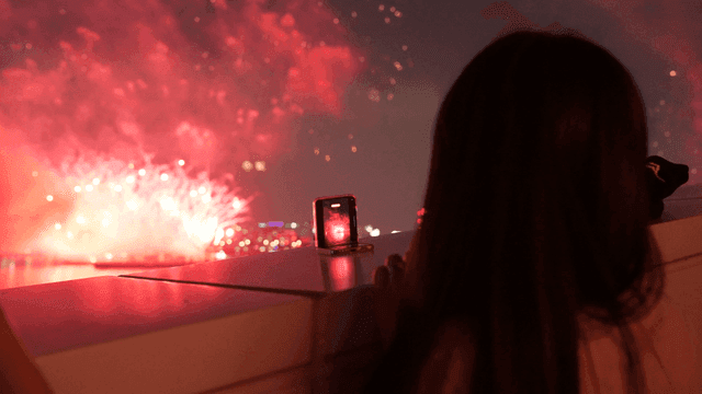 Woman's back as she takes photo of fireworks from her balcony with her cell phone.