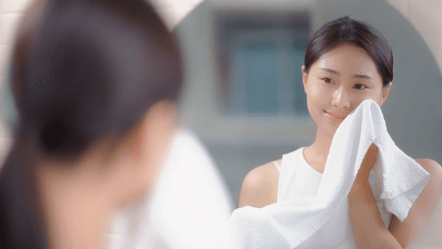 Woman gently drying her face with a towel in front of a bathroom mirror