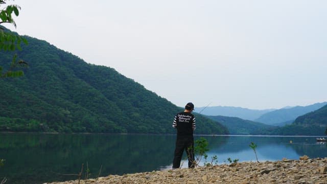 Man fishing by a serene mountain lake on an overcast day