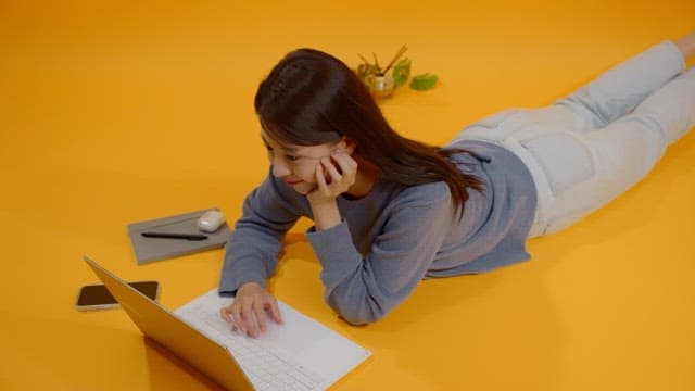 Woman working on her laptop while lying on her stomach in a bright room