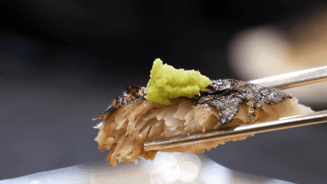 Chopsticks picking up piece of grilled fish topped with wasabi