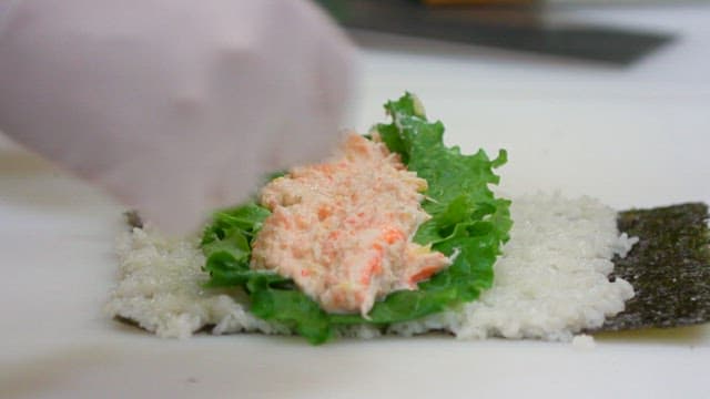 Gloved hand rolling out gimbap with crab meat salad and lettuce on a cutting board