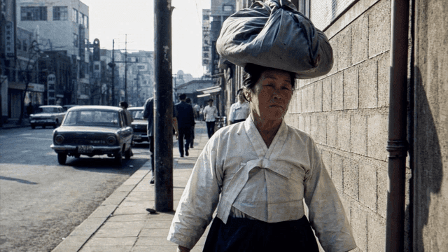 Elderly woman in Hanbok walking down street with load on her head