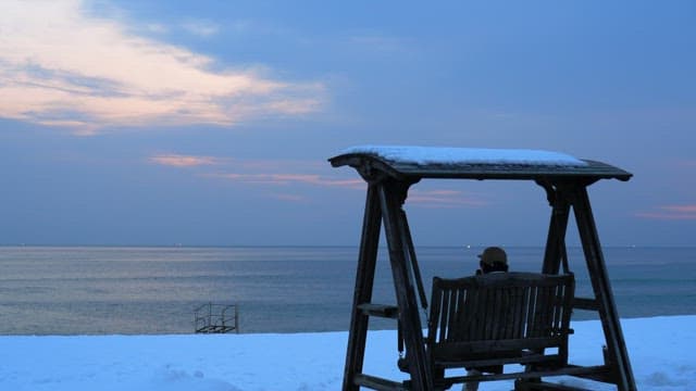 Person sitting on a wooden swing by the snowy beach during sunset